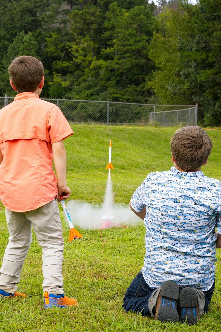 Two kids seen from behind watching a small rocket lift off from a grassy field